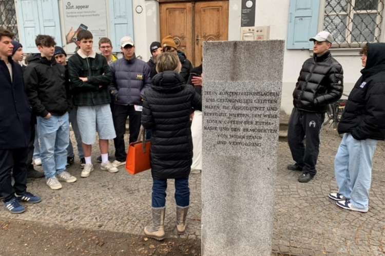 Eine Gruppe Jugendlicher steht um eine Frau, die vor einem Gedenkstein spricht. Die Inschrift erinnert an die Opfer von Konzentrationslagern, Euthanasie und politischer Verfolgung. Die Szene spielt sich vor einem historischen Gebäude mit blauen Fensterläden ab. Die Jugendlichen tragen Freizeitkleidung, einige hören aufmerksam zu, andere haben die Arme verschränkt oder die Hände in den Taschen.